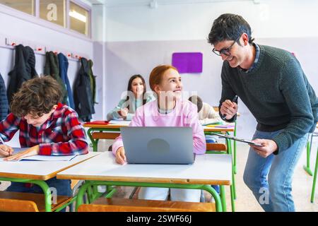 Insegnante sorridente che assiste uno studente utilizzando un laptop in classe, altri studenti che studiano e scrivono alla propria scrivania Foto Stock
