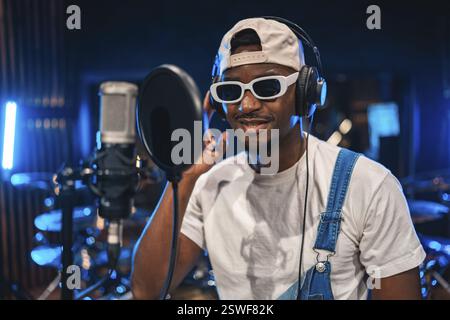 Giovane elegante che registra la voce utilizzando un microfono professionale in uno studio musicale con luci ambiente blu Foto Stock