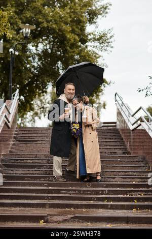 Una coppia amorevole scende i gradini in una giornata di pioggia condividendo un ombrello e bevande calde. Foto Stock