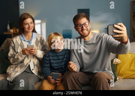 Famiglia di tre persone che si rilassano su un comodo divano con dispositivi, un uomo sorridente con occhiali che reggono lo smartphone, una donna con i capelli lunghi e un ragazzo biondo felice impegnato in attività ricreative Foto Stock