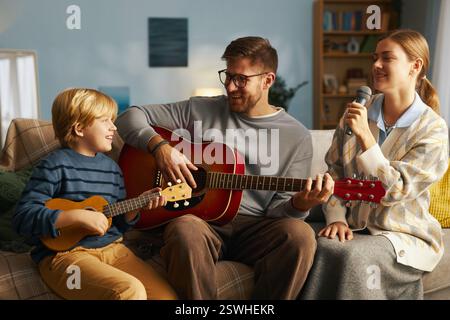 La famiglia si diverte a una sessione musicale con il padre che suona la chitarra, il figlio che suona l'ukulele e la figlia che canta, tutti seduti su un comodo divano, creando un'atmosfera gioiosa Foto Stock