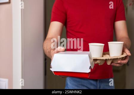 addetto alle consegne in uniforme rossa che consegna al cliente una scatola per alimenti e tazze di carta davanti alla porta d'ingresso Foto Stock