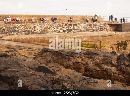 Turisti in piedi e seduti alla fine di New Pier St Ives, Cornovaglia, Inghilterra, Regno Unito, con vista sul mare e sul sole Foto Stock