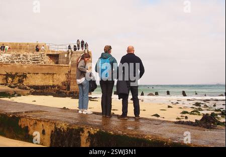 Turisti in piedi sul New Pier e sulla strada rialzata, St Ives, Cornovaglia, Inghilterra, Regno Unito, che si affaccia sul mare sulle onde e su un surfista sul mare Foto Stock