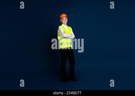 Professionista sicura di sé con giubbotto e elmetto ad alta visibilità, appoggiato su uno sfondo blu scuro Foto Stock