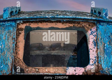 Una finestra rotta sul passato e sul presente: Vista attraverso il vetro avvitato alla cabina di una barca da pesca ridondante e in decomposizione di moderni yacht ormeggiati fuori dal porto di le Fret, un piccolo ex villaggio di pescatori sulla penisola di Crozon a Finistère, Bretagna, Francia. La navigazione a vela e la navigazione da diporto hanno sostituito la pesca come attività principale di le Fret. Foto Stock