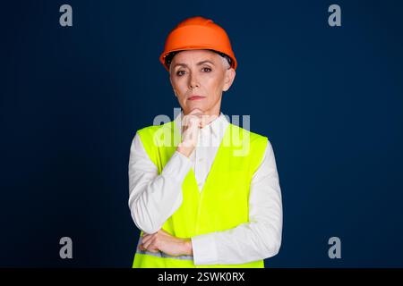 Ingegnere donna maturo in indumenti da lavoro e elmetto ad alta visibilità, che riflette profondamente su uno sfondo blu scuro Foto Stock