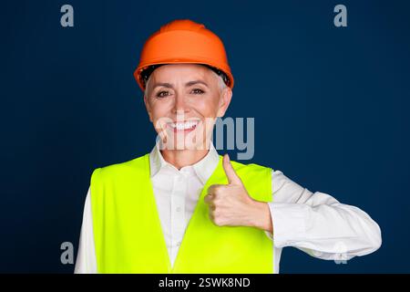 Donna matura e sicura di sé in un outfit da costruzione che regala un pollice su uno sfondo blu scuro Foto Stock