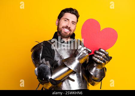 Cavaliere sorridente in un'armatura lucente che regge un cuore rosso vibrante su sfondo giallo brillante che celebra l'amore e il fascino medievale Foto Stock