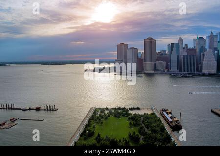 Il vibrante tramonto illumina lo skyline di Manhattan nel centro cittadino mentre le barche scorrono davanti al lussureggiante lungomare Foto Stock