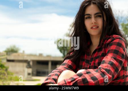 Ritratto di una giovane donna bruna che indossa una camicia rossa e nera, seduta all'aperto con un edificio abbandonato sullo sfondo Foto Stock