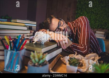 Un giovane stanco in un abbigliamento casual si addormentò alla sua scrivania circondato da pile di libri e documenti in un accogliente ufficio domestico Foto Stock