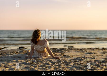 Una giovane donna siede sulla spiaggia sabbiosa, guardando il bellissimo tramonto sulle calme onde dell'oceano. L'atmosfera è tranquilla e serena, perfetta per il re Foto Stock