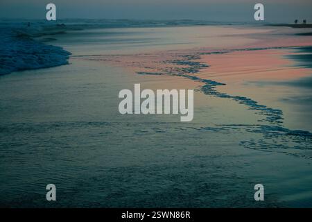 Una tranquilla spiaggia al crepuscolo con onde morbide che si infrangono sulla riva, con un delicato gradiente di riflessi rosa e blu sulla sabbia bagnata Foto Stock