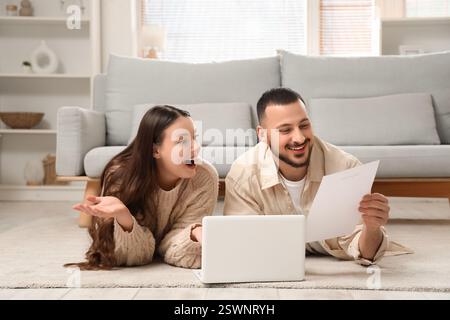 Giovane coppia con modulo di rimborso e laptop sdraiato sul pavimento di casa Foto Stock