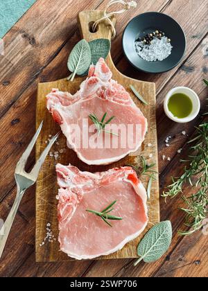 Bistecche di maiale crude sull'osso e erbe aromatiche su un tagliere di legno. Carne di maiale fresca. Direttamente sopra. Foto Stock