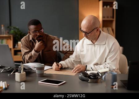 Designer uomo di mezza età con testa rasata che si concentra sul disegno di un'unità di mobili per interni su carta artigianale, mentre collabora con un collega maschio nero alla scrivania in ufficio Foto Stock