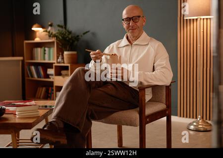 Foto ritratto di un dipendente maschio sorridente con la testa rasata seduta in poltrona e gustando un pranzo veloce e bilanciato che l'uomo ha ordinato per la consegna al proprio ufficio Foto Stock