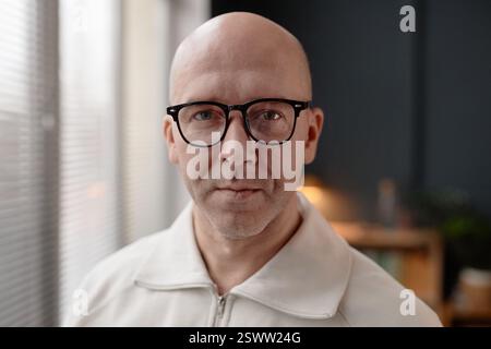 Ritratto di un uomo di mezza età sicuro con la testa rasata che indossa gli occhiali che guarda la fotocamera mentre è in piedi alla luce del giorno che proietta attraverso la finestra dell'ufficio domestico, copia lo spazio Foto Stock