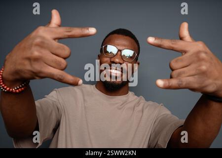 Inquadratura di un allegro uomo Nero che sorride mentre fa segno di corna che esprimono energia positiva in posa in studio su sfondo grigio Foto Stock