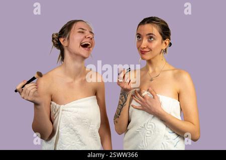 Due giovani donne con asciugamani avvolti intorno al corpo stanno applicando il trucco, una sta ridendo e tiene in mano una spazzola, l'altra un rossetto Foto Stock