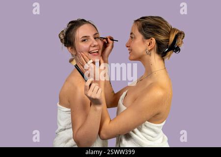 Due amici si divertono ad applicare il trucco insieme, avvolti in asciugamani dopo la doccia, godendosi la loro routine di benessere Foto Stock