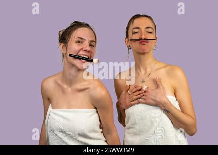 Due amiche giocherellone si stanno godendo una giornata in spa, indossando asciugamani e facendosi facce divertenti con le spazzole per il trucco Foto Stock