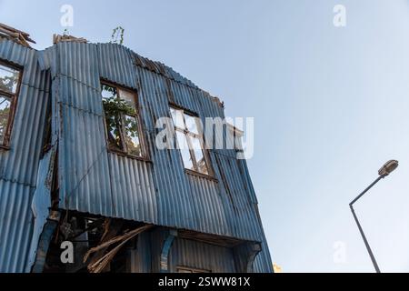 Un edificio fatiscente rivestito di pannelli di metallo ondulato blu, con finestre rotte e fogliame verde che cresce all'interno. Foto Stock