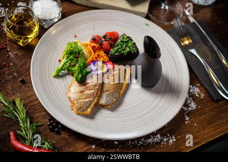 Filetto di spigola grigliato con broccoli, pomodori ciliegini, nastri di carote e fiori commestibili. Servito con un contorno di erbe fresche e una brocca con salsa scura Foto Stock