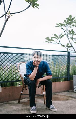 Un giovane asiatico siede pensiamente su una sedia su una terrazza sul tetto. Sembra premuroso e rilassato, godendo di un momento di pace solitudine in mezzo al Foto Stock