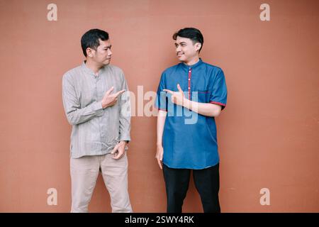 Due giovani uomini con espressioni amichevoli che si puntano l'un l'altro mentre si alzano contro un muro color terracotta. Sono vestiti con abbigliamento casual. Foto Stock