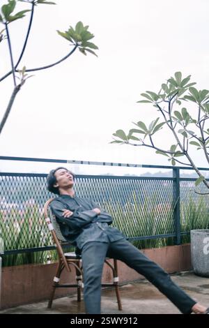 Un uomo asiatico è seduto su una sedia sulla terrazza e si rilassa. Ha un aspetto premuroso mentre contempla la vita, in un ambiente tranquillo. E' casualmente il dottor Foto Stock