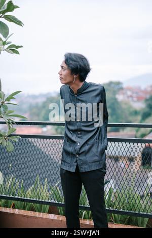 Un giovane uomo elegante guarda con cura su un paesaggio verde da una terrazza sul tetto. È vestito di nero e sembra contemplare la S Foto Stock