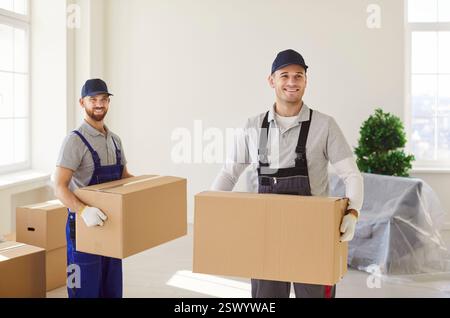 Giovani uomini che si muovono in uniforme tenendo una scatola di cartone in piedi in un nuovo appartamento vuoto il giorno del trasloco Foto Stock