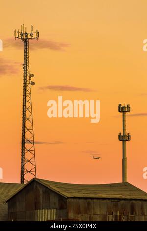 Torre con antenna per telefono cellulare sulla parte superiore. Un aereo che vola nel cielo. Il cielo è arancione e il sole sta tramontando Foto Stock