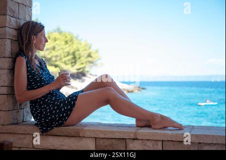 Una donna con un abito estivo si appoggia contro un muro di pietra, tenendo in mano una tazza di caffè o tè mentre osserva l'oceano blu, godendo di una tranquilla vista costiera. Foto Stock
