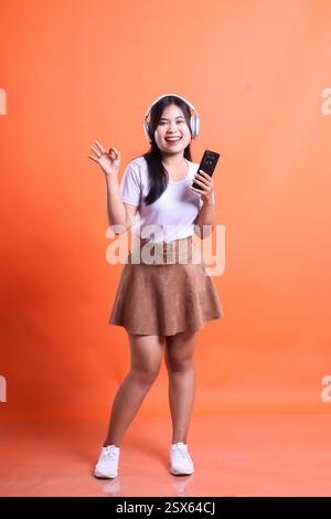 Una donna asiatica ascolta la musica con le cuffie e tiene in mano uno smartphone sorridente. Indossa una gonna e una camicia, isolata sul retro arancione Foto Stock