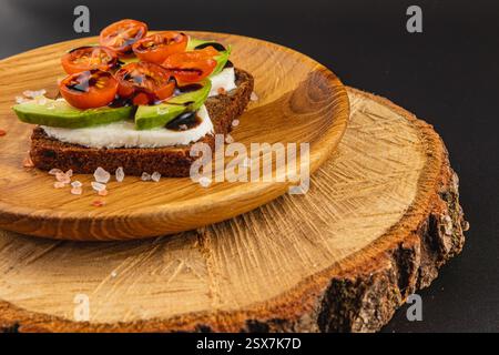 Un delizioso panino a faccia aperta presenta una cremosa spalmazione condita con avocado a fette, pomodori ciliegini e glassa balsamica, servita su un piatto rustico di legno Foto Stock