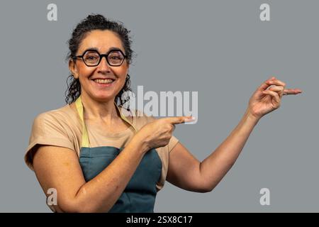 Donna matura felice che indossa grembiule e occhiali puntando da parte con le dita indice e mostrando qualcosa su sfondo grigio Foto Stock
