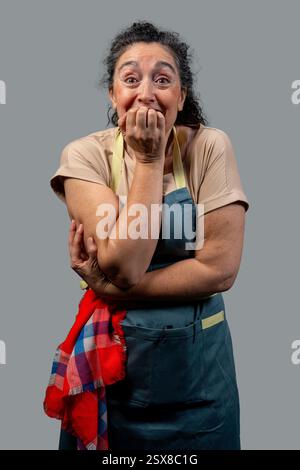 Chef donna anziana con grembiule e asciugamano da cucina rosso e blu che le mordono le unghie, mostrando ansia e nervosismo su sfondo grigio Foto Stock
