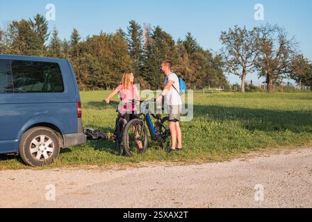 Il giovane uomo e una donna si preparano per la bicicletta fuoristrada, abbattendo mountain bike elettriche dal portabiciclette sul camper. Foto Stock