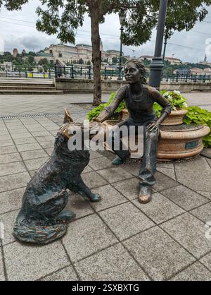Ragazza con la sua statua del cane a Budapest, una scultura emozionante lungo la passeggiata sul Danubio, con uno splendido sfondo del castello di Buda. Foto Stock