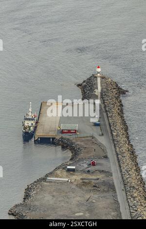 Una nave ormeggiata in un molo con un faro, adagiata su una tranquilla costa rocciosa sotto un cielo coperto, montagne, rocce, strada, mare, oceano, acqua, natur Foto Stock