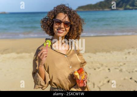 Ritratto di una giovane donna araba ciuta con capelli ricci e occhiali da sole che mangiano frutta tropicale a fette in spiaggia Foto Stock