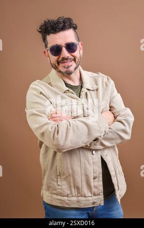 Ritratto di un uomo sorridente e sicuro di sé che indossa occhiali da sole e una giacca in denim beige in piedi con le braccia incrociate su sfondo marrone Foto Stock