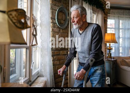 Vecchio con la stampella che si sente deluso, perso in pensieri tristi da solo a casa. Vedova uomo maturo. Foto Stock