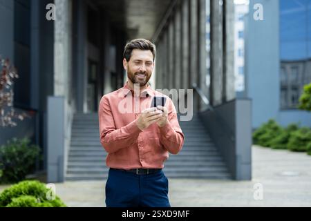 Uomo d'affari di successo fuori dall'edificio di uffici con il telefono in mano, uomo che digita un messaggio di testo e utilizza l'app online mentre cammini in città. Foto Stock