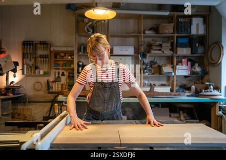 Falegname donna in occhiali protettivi taglia pezzi di legno su macchine per la lavorazione del legno in officina artigianale Foto Stock