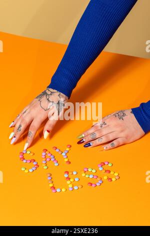 Una composizione giocosa con "SAY LESS" impreziosita da caramelle colorate su una superficie arancione, con mani tatuate e unghie vivaci che aggiungono contrasto. Foto Stock