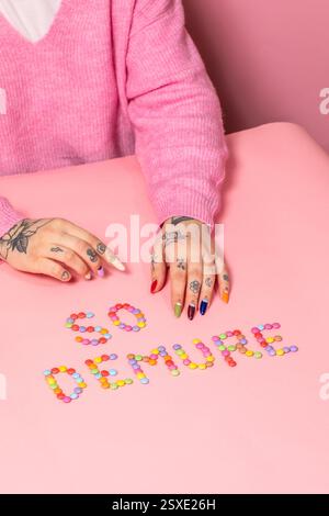 Una composizione giocosa con "SO DEMURE" impreziosita da colorate caramelle su una superficie rosa, con mani tatuate e unghie vivaci che aggiungono contrasto. Foto Stock